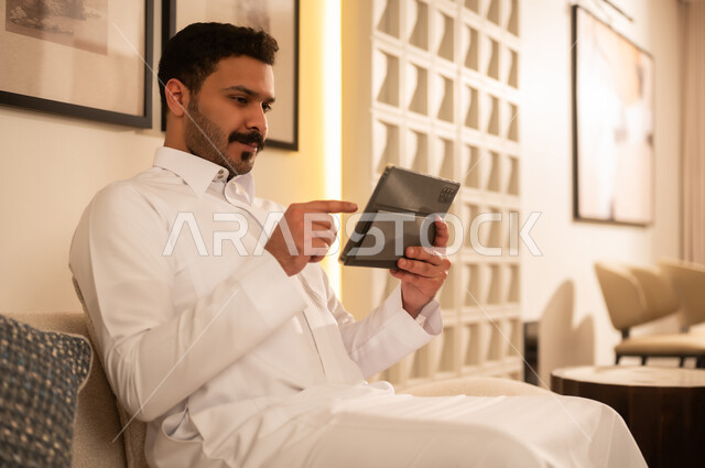 رجل سعودي بالثوب الأبيض يستخدم جهاز لوحي في المنزل