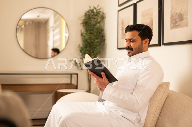 رجل سعودي بالثوب التقليدي يقرأ كتاباً في منزل عصري