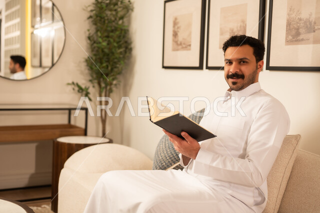 رجل سعودي بالثوب الأبيض يقرأ كتاباً في غرفة معيشة حديثة
