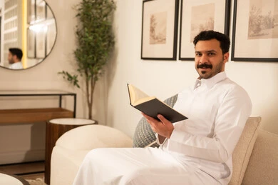 رجل سعودي بالثوب الأبيض يقرأ كتاباً في غرفة معيشة حديثة