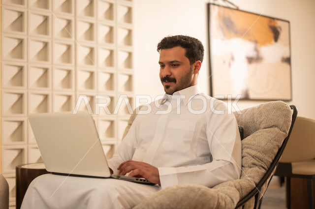 رجل سعودي بالثوب يستخدم لابتوب في غرفة معيشة عصرية
