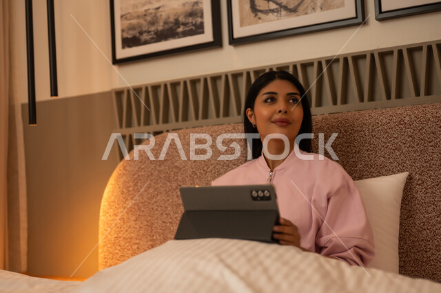امرأة عربية تستخدم جهازاً لوحياً في غرفة نوم حديثة ليلاً