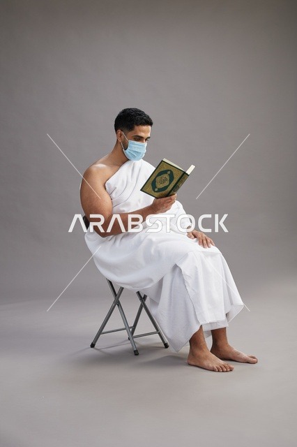 بورتريه لرجل عربي  سعودي  خليجي بملابس الإحرام يجلس على كرسي يقرأ القرآن ويرتدي كمامة, الوقايه من فايروس كورونا, خلفيه رماديه