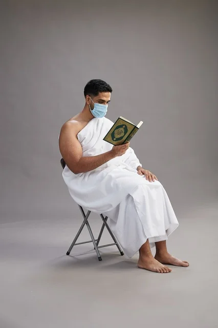 A portrait of an Arab Saudi Gulf man in Ihram clothing sitting on a chair reading the Quran and wearing a mask for protection against the coronavirus, with a gray background.