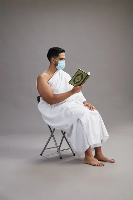 A portrait of an Arab Saudi Gulf man in Ihram clothing sitting on a chair reading the Quran and wearing a mask for protection against the coronavirus, with a gray background.