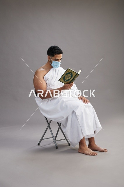بورتريه لرجل عربي  سعودي  خليجي بملابس الإحرام يجلس على كرسي يقرأ القرآن ويرتدي كمامة, الوقايه من فايروس كورونا, خلفيه رماديه