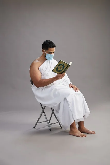 A portrait of an Arab Saudi Gulf man in Ihram clothing sitting on a chair reading the Quran and wearing a mask for protection against the coronavirus, with a gray background.