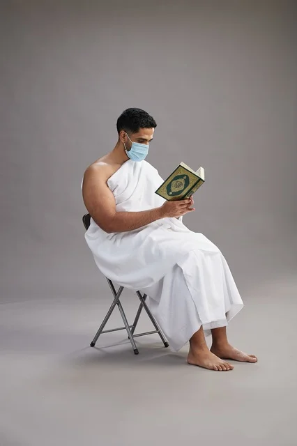 A portrait of an Arab Saudi Gulf man in Ihram clothing sitting on a chair reading the Quran and wearing a mask for protection against the coronavirus, with a gray background.