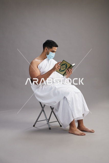 بورتريه لرجل عربي  سعودي  خليجي بملابس الإحرام يجلس على كرسي يقرأ القرآن ويرتدي كمامة, الوقايه من فايروس كورونا, خلفيه رماديه
