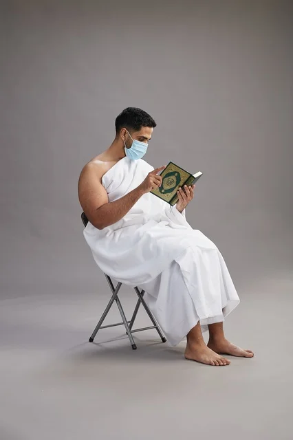 A portrait of an Arab Saudi Gulf man in Ihram clothing sitting on a chair reading the Quran and wearing a mask for protection against the coronavirus, with a gray background.