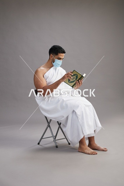 بورتريه لرجل عربي  سعودي  خليجي بملابس الإحرام يجلس على كرسي يقرأ القرآن ويرتدي كمامة, الوقايه من فايروس كورونا, خلفيه رماديه
