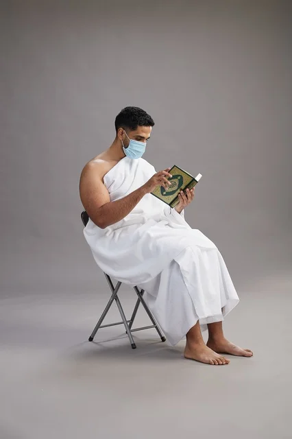 A portrait of an Arab Saudi Gulf man in Ihram clothing sitting on a chair reading the Quran and wearing a mask for protection against the coronavirus, with a gray background.