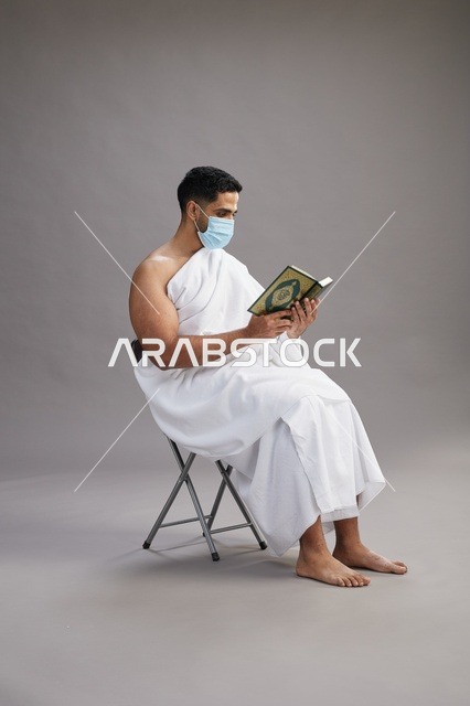 بورتريه لرجل عربي  سعودي  خليجي بملابس الإحرام يجلس على كرسي يقرأ القرآن ويرتدي كمامة, الوقايه من فايروس كورونا, خلفيه رماديه