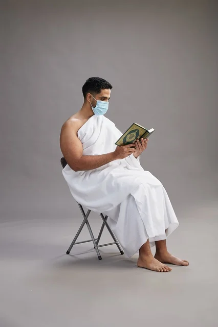 A portrait of an Arab Saudi Gulf man in Ihram clothing sitting on a chair reading the Quran and wearing a mask for protection against the coronavirus, with a gray background.