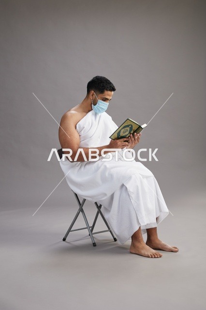 بورتريه لرجل عربي  سعودي  خليجي بملابس الإحرام يجلس على كرسي يقرأ القرآن ويرتدي كمامة, الوقايه من فايروس كورونا, خلفيه رماديه