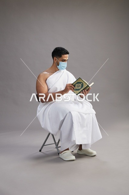 بورتريه لرجل عربي  سعودي  خليجي بملابس الإحرام يجلس على كرسي يقرأ القرآن ويرتدي كمامة, الوقايه من فايروس كورونا, خلفيه رماديه