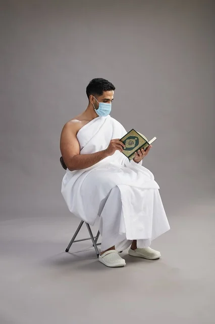 A portrait of an Arab Saudi Gulf man in Ihram clothing sitting on a chair reading the Quran and wearing a mask for protection against the coronavirus, with a gray background.