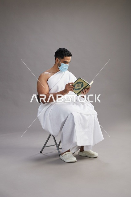 بورتريه لرجل عربي  سعودي  خليجي بملابس الإحرام يجلس على كرسي يقرأ القرآن ويرتدي كمامة, الوقايه من فايروس كورونا, خلفيه رماديه