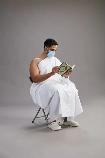 A portrait of an Arab Saudi Gulf man in Ihram clothing sitting on a chair reading the Quran and wearing a mask for protection against the coronavirus, with a gray background.