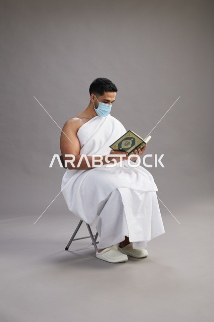 بورتريه لرجل عربي  سعودي  خليجي بملابس الإحرام يجلس على كرسي يقرأ القرآن ويرتدي كمامة, الوقايه من فايروس كورونا, خلفيه رماديه