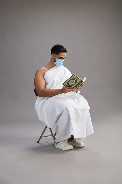 A portrait of an Arab Saudi Gulf man in Ihram clothing sitting on a chair reading the Quran and wearing a mask for protection against the coronavirus, with a gray background.
