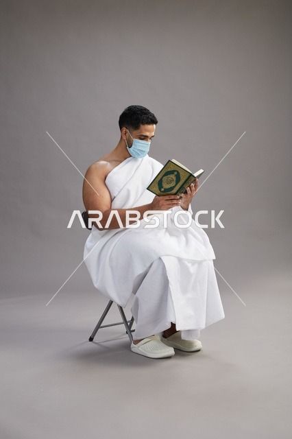 بورتريه لرجل عربي  سعودي  خليجي بملابس الإحرام يجلس على كرسي يقرأ القرآن ويرتدي كمامة, الوقايه من فايروس كورونا, خلفيه رماديه