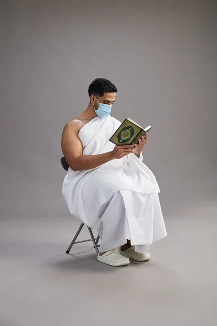 A portrait of an Arab Saudi Gulf man in Ihram clothing sitting on a chair reading the Quran and wearing a mask for protection against the coronavirus, with a gray background.