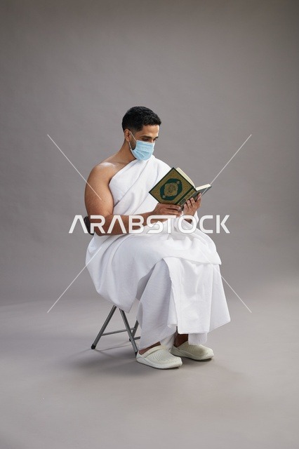 بورتريه لرجل عربي  سعودي  خليجي بملابس الإحرام يجلس على كرسي يقرأ القرآن ويرتدي كمامة, الوقايه من فايروس كورونا, خلفيه رماديه