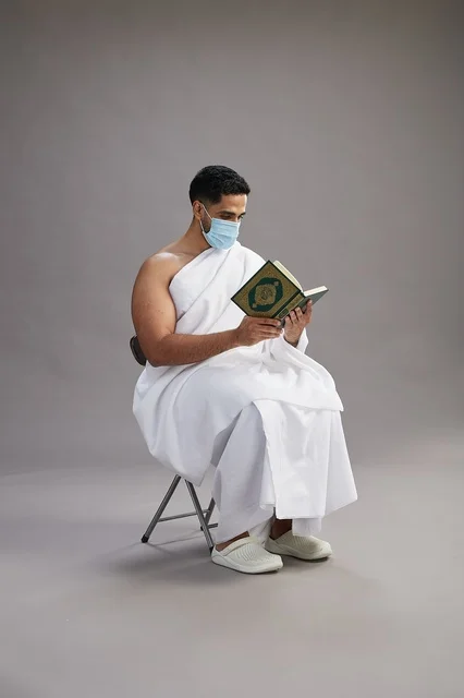 A portrait of an Arab Saudi Gulf man in Ihram clothing sitting on a chair reading the Quran and wearing a mask for protection against the coronavirus, with a gray background.