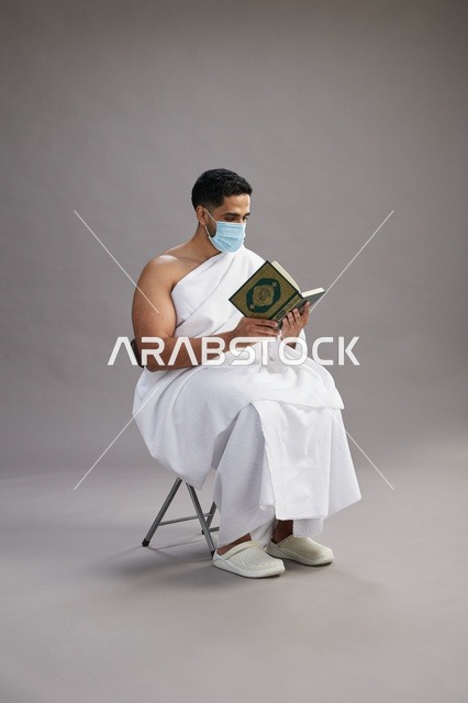بورتريه لرجل عربي  سعودي  خليجي بملابس الإحرام يجلس على كرسي يقرأ القرآن ويرتدي كمامة, الوقايه من فايروس كورونا, خلفيه رماديه