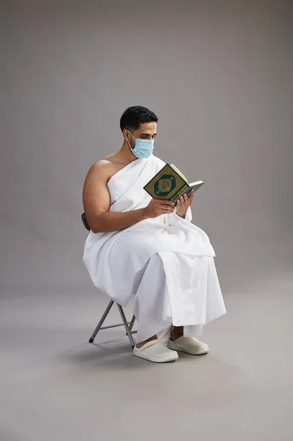 A portrait of an Arab Saudi Gulf man in Ihram clothing sitting on a chair reading the Quran and wearing a mask for protection against the coronavirus, with a gray background.