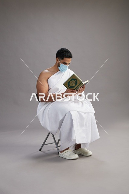بورتريه لرجل عربي  سعودي  خليجي بملابس الإحرام يجلس على كرسي يقرأ القرآن ويرتدي كمامة, الوقايه من فايروس كورونا, خلفيه رماديه