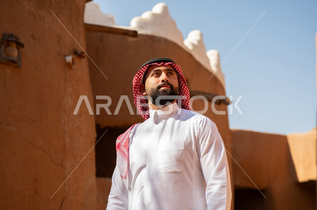 A Saudi Arabian Gulf Arab man wearing traditional attire, a white thobe and a shemagh, looks at something above. It is the anniversary of the establishment of the first Saudi state by Imam Muhammad bin Saud, which began on February 22, 1727. The historical and cultural depth of the Kingdom, national celebrations in the central region.