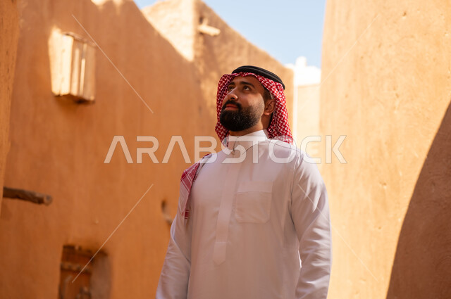 A Saudi Arabian Gulf Arab man wearing traditional attire, a white thobe and a shemagh, looks at something above. It is the anniversary of the establishment of the first Saudi state by Imam Muhammad bin Saud, which began on February 22, 1727. The historical and cultural depth of the Kingdom, national celebrations in the central region.