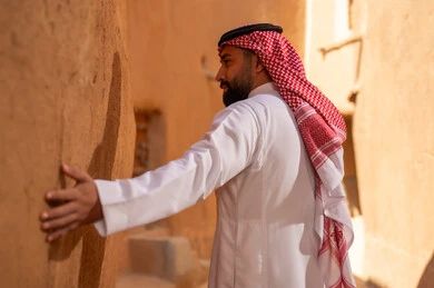 A side view of a Saudi Arabian Gulf man wearing traditional attire, a white thobe and a shemagh, with his hand on a clay wall, commemorating the founding of the first Saudi state by Imam Muhammad bin Saud, on the day we began, February 22, 1727 AD, the historical and cultural depth of the Kingdom, national celebrations in the central region.
