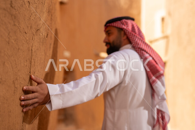 صورة من الجانب لرجل عربي سعودي خليجي يرتدي الزي التقليدي ثوب أبيض وشماخ يده على جدار طيني، ذكرى تأسيس الدوله السعوديه الاولى على يد الامام محمد بن سعود, يوم بدينا 22 فبراير 1727م, العمق التاريخي والحضاري للمملكة،الإحتفالات الوطنيه في المنطقه الوسطى