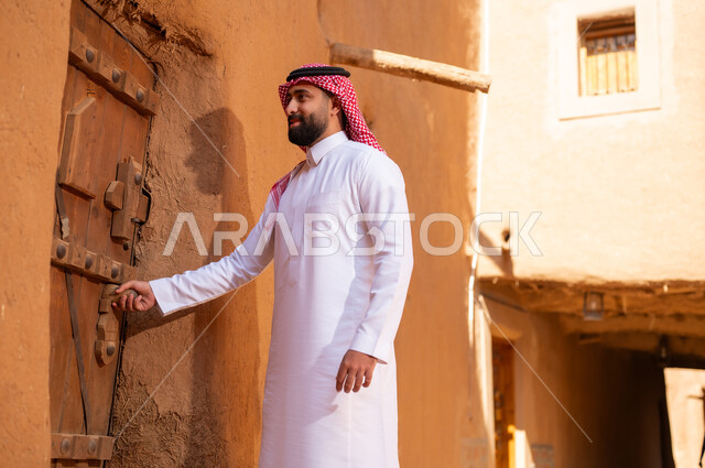 رجل عربي سعودي خليجي يرتدي الزي التقليدي ثوب أبيض وشماخ ويمسك باب خشبي، ذكرى تأسيس الدوله السعوديه الاولى على يد الامام محمد بن سعود, يوم بدينا 22 فبراير 1727م, العمق التاريخي والحضاري للمملكة،الإحتفالات الوطنيه في المنطقه الوسطى