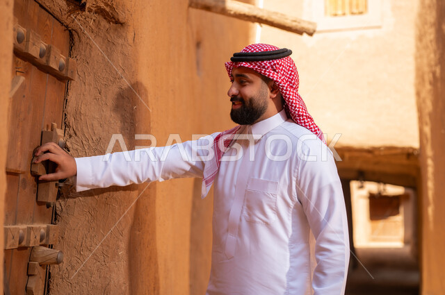 An Arab Saudi Gulf man wearing traditional attire, a white thobe, holding a wooden door, commemorating the founding of the first Saudi state by Imam Muhammad bin Saud, on the day we began, February 22, 1727 AD, the historical and cultural depth of the Kingdom, national celebrations in the central region.
