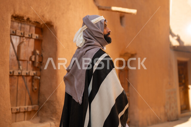 صورة من الخلف رجل عربي سعودي خليجي يرتدي زي يوم التأسيسي يقف أمام مبنى طيني وينظر الى شي ما في الأعلى، ذكرى تأسيس الدوله السعوديه الاولى على يد الامام محمد بن سعود, يوم بدينا 22 فبراير 1727م, العمق التاريخي والحضاري للمملكة،الإحتفالات الوطنيه في المنطقه الوسطى