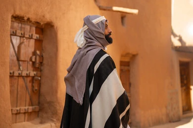 A picture from behind of an Arab Saudi Gulf man wearing the founding day attire, standing in front of a mud building and looking at something above, commemorating the founding of the first Saudi state by Imam Muhammad bin Saud, on the day we began, February 22, 1727 AD, the historical and cultural depth of the Kingdom, national celebrations in the central region.
