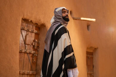 صورة من الخلف رجل عربي سعودي خليجي يرتدي زي يوم التأسيسي يقف أمام مبنى طيني وينظر الى شي ما في الأعلى، ذكرى تأسيس الدوله السعوديه الاولى على يد الامام محمد بن سعود, يوم بدينا 22 فبراير 1727م, العمق التاريخي والحضاري للمملكة،الإحتفالات الوطنيه في المنطقه الوسطى