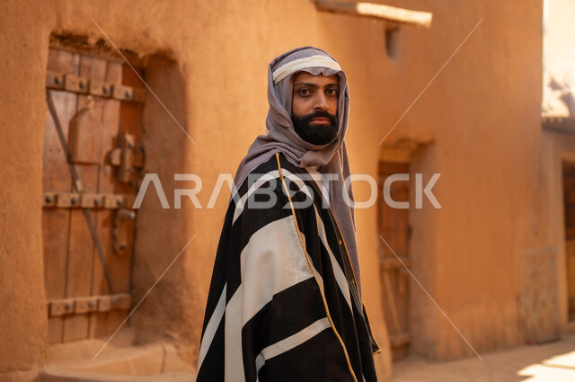 رجل عربي سعودي خليجي يرتدي زي يوم التأسيسي يقف أمام مبنى طيني وينظر الى شي ما بفخر، ذكرى تأسيس الدوله السعوديه الاولى على يد الامام محمد بن سعود, يوم بدينا 22 فبراير 1727م, العمق التاريخي والحضاري للمملكة، هوية اليوم الوطني السعودي 92