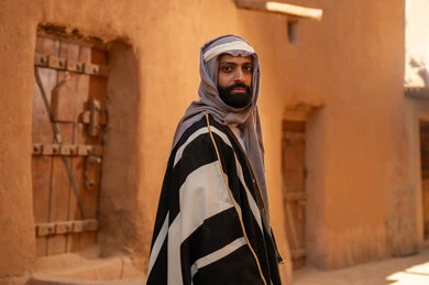A Saudi Arabian Gulf Arab man wearing the founding day attire stands in front of a mud building, looking at something with pride, commemorating the establishment of the first Saudi state by Imam Muhammad bin Saud, on the day we began, February 22, 1727 AD, the historical and cultural depth of the Kingdom, the identity of Saudi National Day 92.