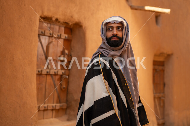 A Saudi Arabian Gulf Arab man wearing the founding day attire stands in front of a mud building, looking at something with pride, commemorating the establishment of the first Saudi state by Imam Muhammad bin Saud, on the day we began, February 22, 1727 AD, the historical and cultural depth of the Kingdom, the identity of Saudi National Day 92.