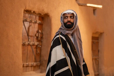 A Saudi Arabian Gulf Arab man wearing the founding day attire stands in front of a mud building, looking at something with pride, commemorating the establishment of the first Saudi state by Imam Muhammad bin Saud, on the day we began, February 22, 1727 AD, the historical and cultural depth of the Kingdom, the identity of Saudi National Day 92.