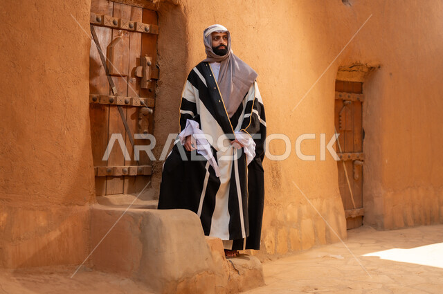 An Arab Saudi Gulf man wearing the founding day attire stands in front of the door of a mud building, commemorating the establishment of the first Saudi state by Imam Muhammad bin Saud, on the day we began, February 22, 1727 AD, the historical and cultural depth of the Kingdom, the identity of Saudi National Day 92.
