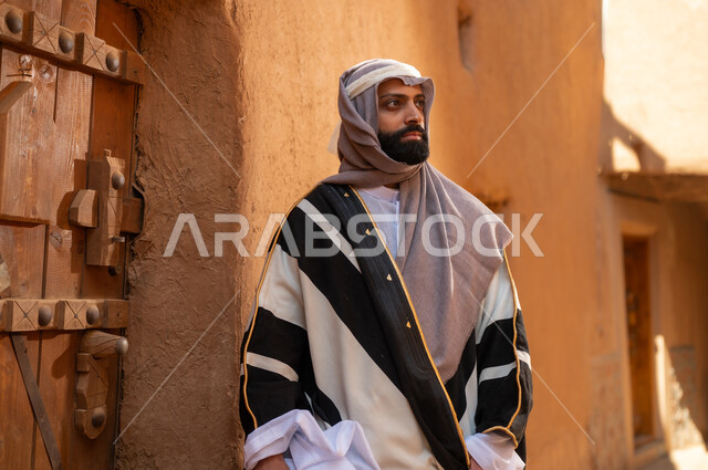 رجل عربي سعودي خليجي يرتدي زي يوم التأسيسي يقف أمام باب مبنى طيني، ذكرى تأسيس الدوله السعوديه الاولى على يد الامام محمد بن سعود, يوم بدينا 22 فبراير 1727م, العمق التاريخي والحضاري للمملكة، هوية اليوم الوطني السعودي 92