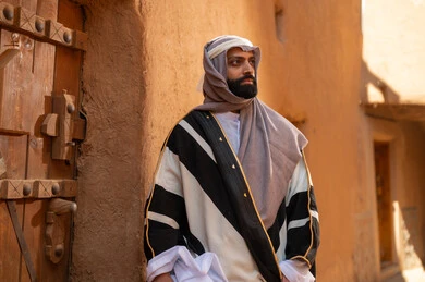 رجل عربي سعودي خليجي يرتدي زي يوم التأسيسي يقف أمام باب مبنى طيني، ذكرى تأسيس الدوله السعوديه الاولى على يد الامام محمد بن سعود, يوم بدينا 22 فبراير 1727م, العمق التاريخي والحضاري للمملكة، هوية اليوم الوطني السعودي 92