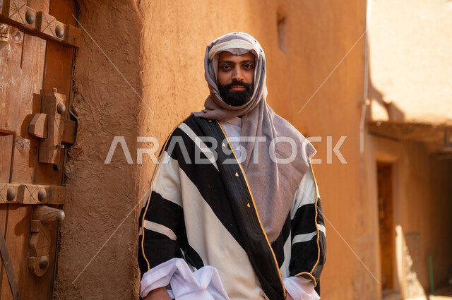 رجل عربي سعودي خليجي يرتدي زي يوم التأسيسي يقف أمام باب مبنى طيني، ذكرى تأسيس الدوله السعوديه الاولى على يد الامام محمد بن سعود, يوم بدينا 22 فبراير 1727م, العمق التاريخي والحضاري للمملكة، هوية اليوم الوطني السعودي 92