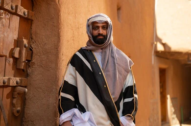 An Arab Saudi Gulf man wearing the founding day attire stands in front of the door of a mud building, commemorating the establishment of the first Saudi state by Imam Muhammad bin Saud, on the day we began, February 22, 1727 AD, the historical and cultural depth of the Kingdom, the identity of Saudi National Day 92.